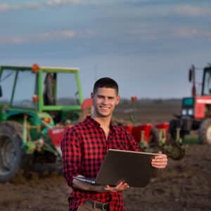 Young farmer with laptop standing in field with tractors in background
